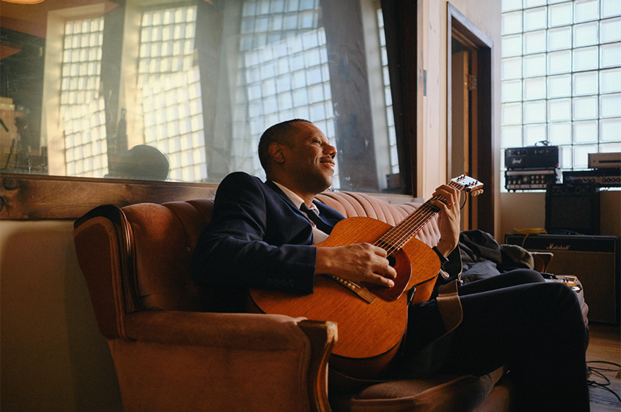 A man playing the guitar sat on a sofa.