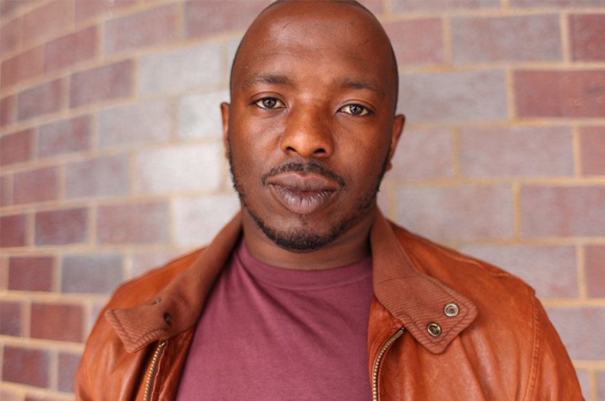 A photo of Nick Makoha standing in front of a brick wall.