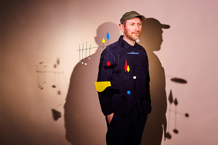 A man wearing a cap with his hands in his pocket with a few red, yellow and blue shapes floating in front of him.