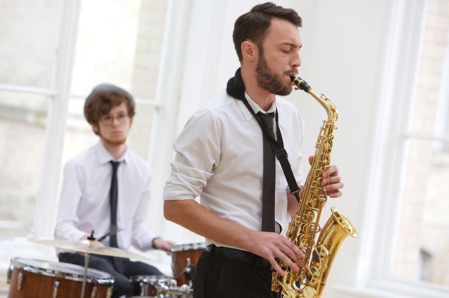 Male student playing a saxophone with another male student playing a drumkit behind.