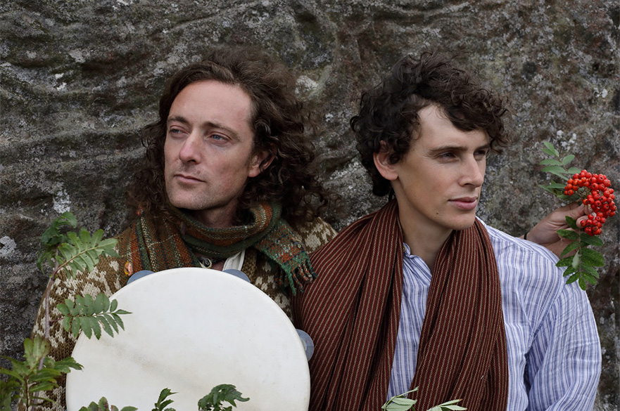 Two men stood in front of an rocky wall with a few branches surrounding them