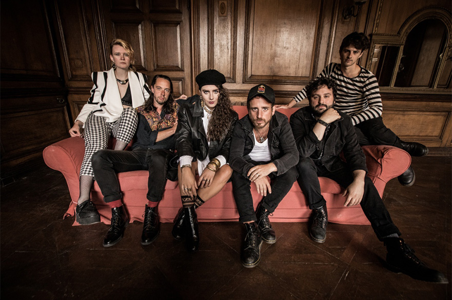 A band of six people sat on a red sofa, all looking at the camera.