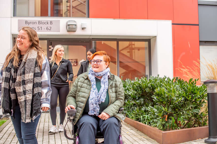 Four students leaving Byron Block B and heading out into the city. In the foreground is a student in an electric wheelchair smiling with a fellow student.