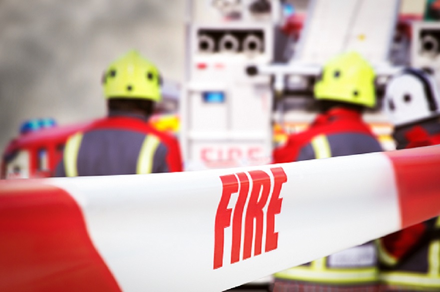 Scene of incident is cordoned off with red and white tape, reading ‘fire’. Out of focus in the background are three firefighters, two fire engine appliances and smoke billowing through the air