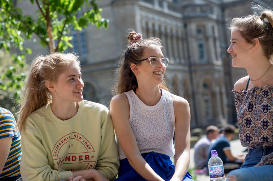Three female students sat outside the Newton building in the sun