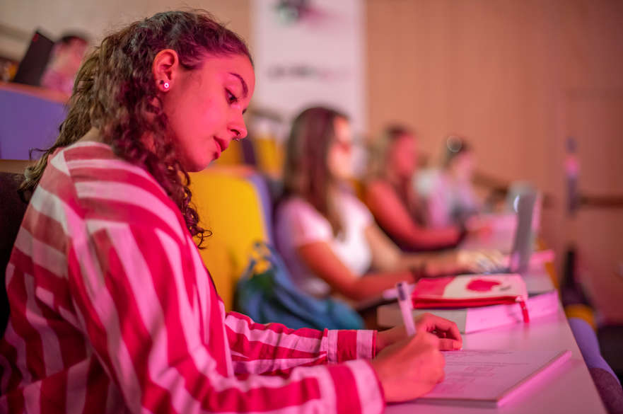 A student in a stripy red and white shirt writing notes in a lecture theatre.