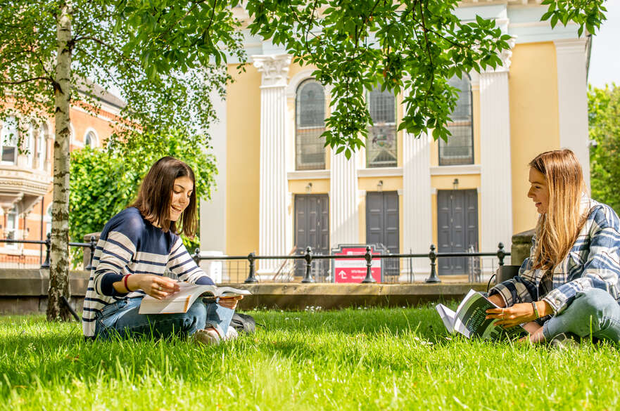 Two students reading textbooks on the grass outside our Arkwright building. University Hall is visible in the background.