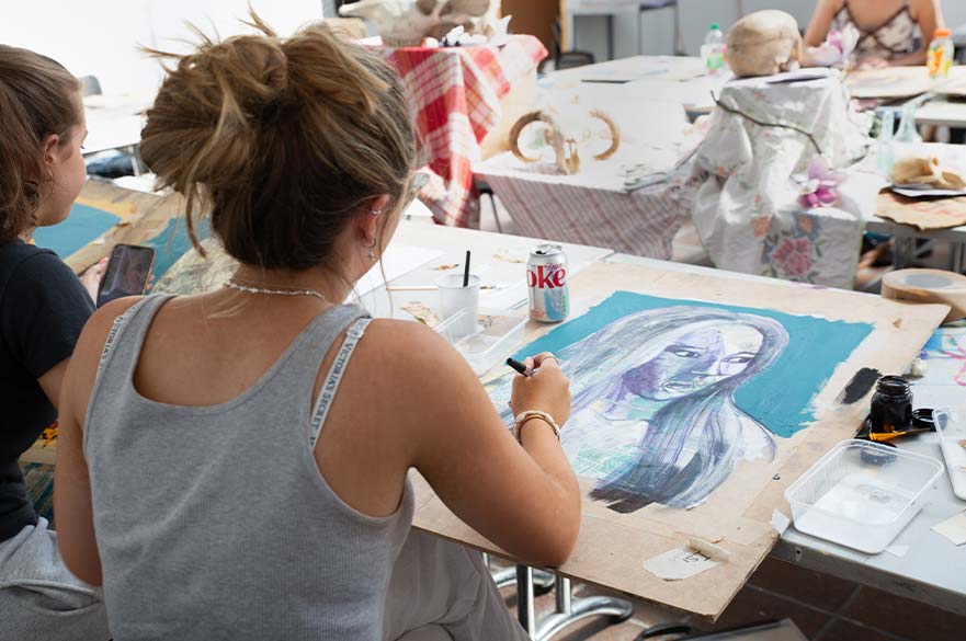 A girl sat in a large art studio, shown from behind to show the sketchbook she's working on