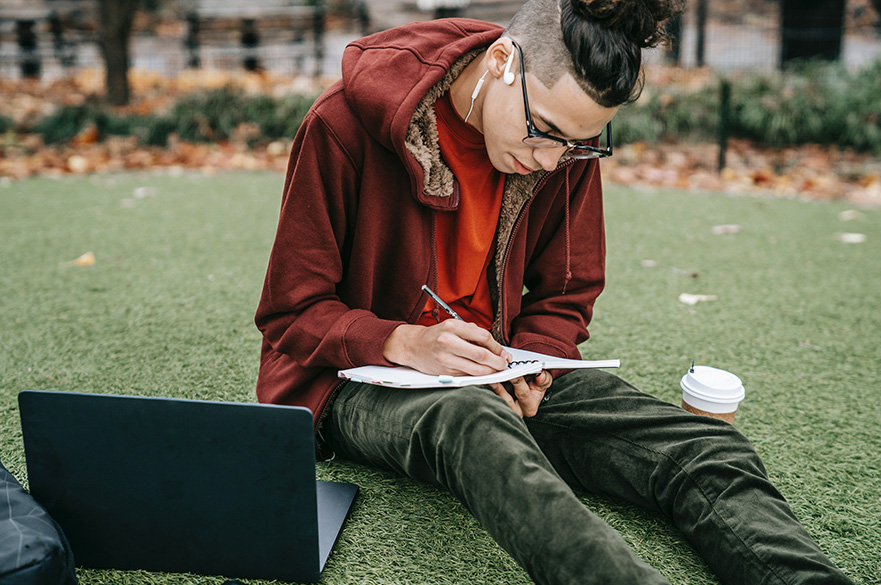 A young man sitting in a relaxed position outdoors, leaning over a notebook and writing. He is wearing a red hoodie and green jeans. A laptop and coffee cup are beside him on the grass.