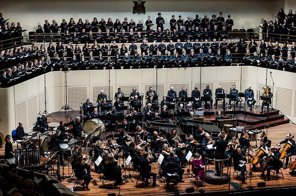 A huge symphony orchestra on a large stage with a large choir singing above the orchestra in high choir stalls.