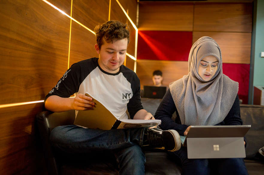 Two students sat side by side, one is a white man and one is a woman wearing a hijab.