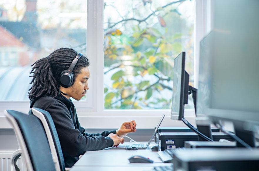 A student with dreadlocks concentrating on a laptop and monitor at a desk bank beside the window.