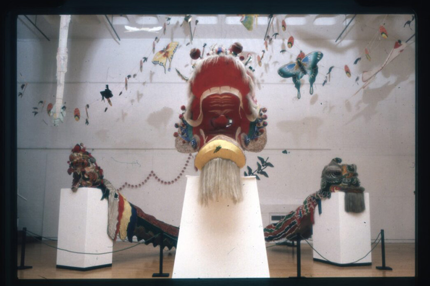 An art installation featuring three colorful traditional Chinese dragon or lion dance heads mounted on white pedestals in a gallery space.