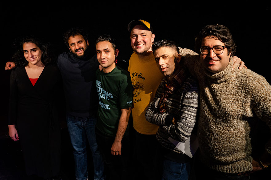 Six comedians with their arms around each other, standing in front of a black background.