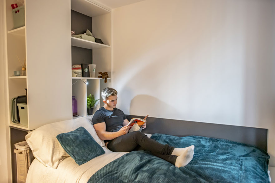 A student sat reading on his blue halls of residence bed. Behind him are neatly organised shelves.