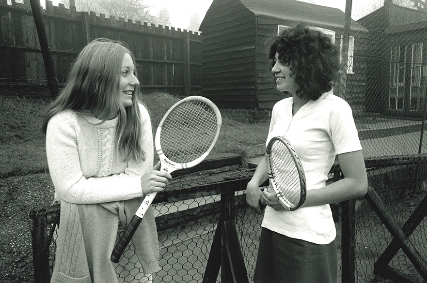 Two girls talking at a tennis net.
