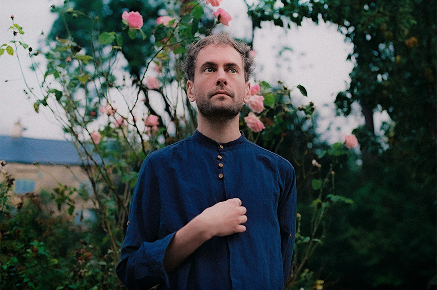 A man standing in front of a pink rose bush with his hand in front of his chest.
