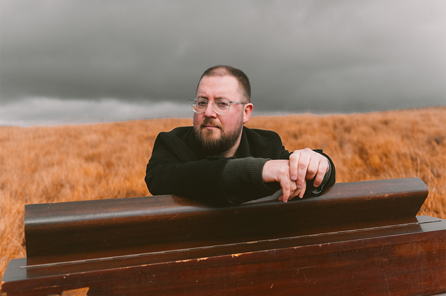A man with glasses leaning his arms over the top of a small piano in wheat field.