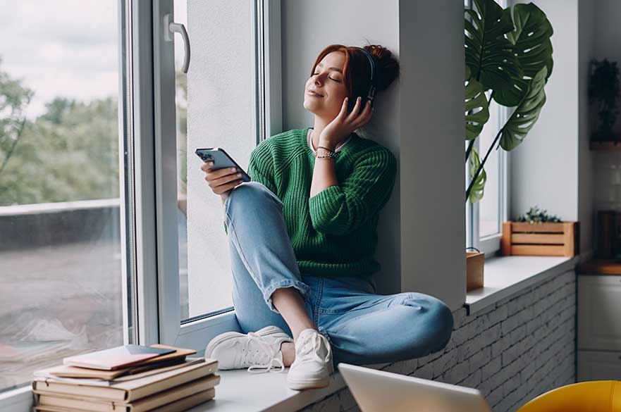 Woman sitting on a windowsill with headphones on and phone in her hands with eyes closed