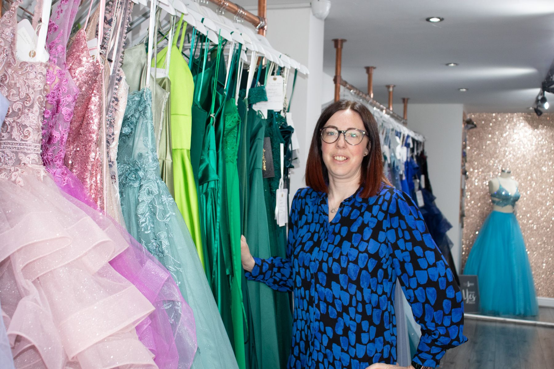 A lady with dark brown hair and glasses is stood next to a rail of different coloured sparkly prom dresses