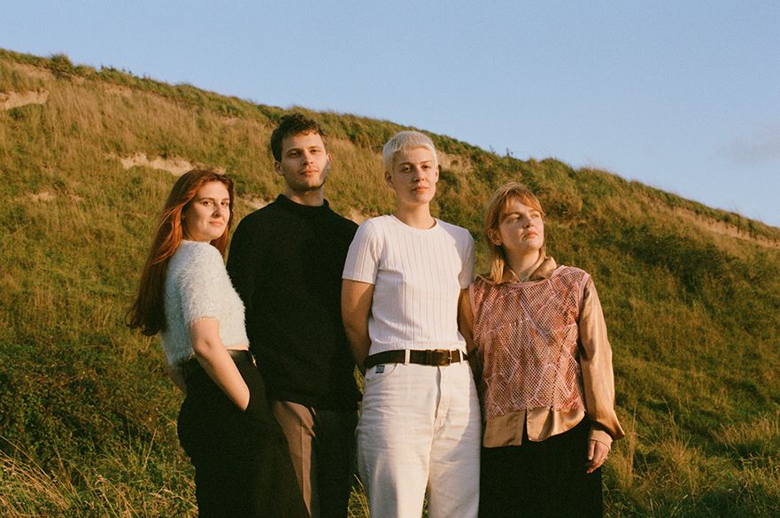 Four young people stand together in a rural setting, against a backdrop of a grassy hill. One has long red hair, another has a blonde pixie cut and a third has shoulder-length strawberry hair. The fourth is a man with short brown hair.