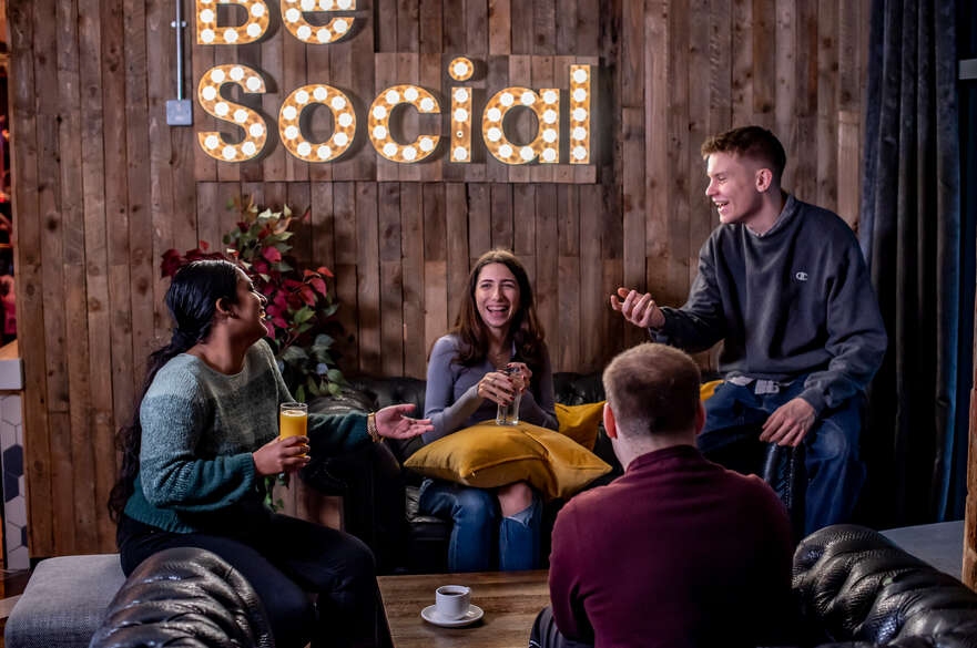 Four students sitting on couches laugh together under an illuminated sign spelling out the words 