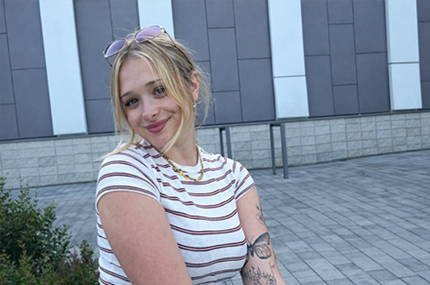 A person taking a selfie photograph, posing in front of their camera, smiling, standing outside a building.