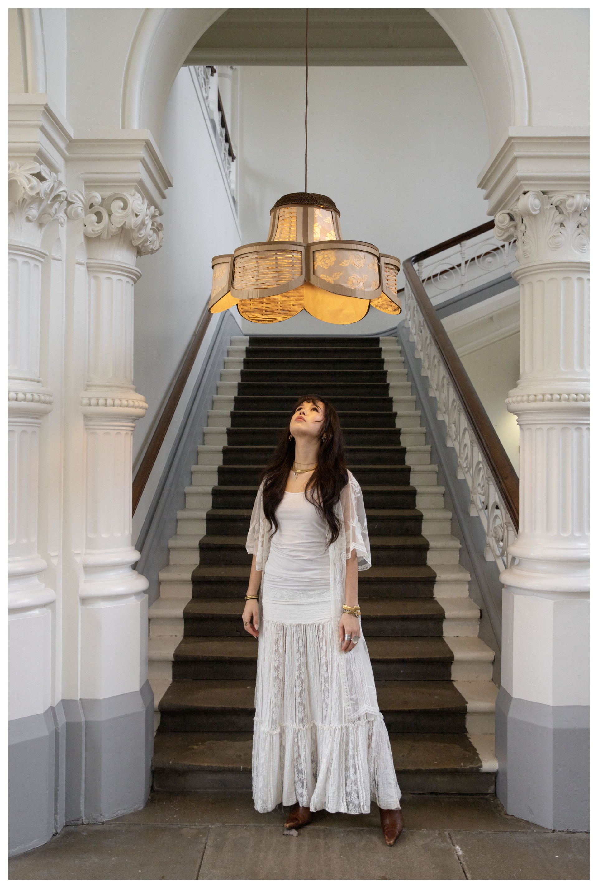 A person standing at the foot of some stairs looking up at a light fitting with lampshade.