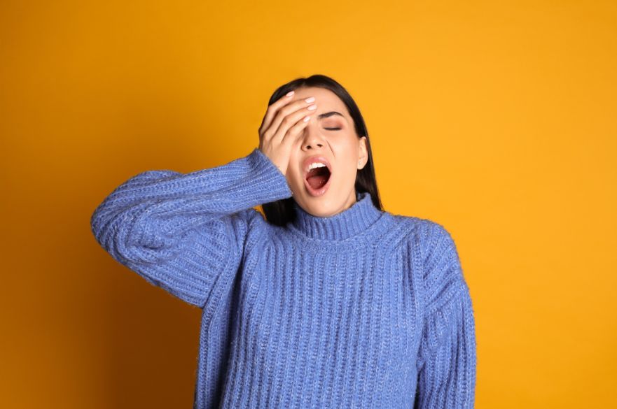 Woman in a blue jumper yawning against a yellow background