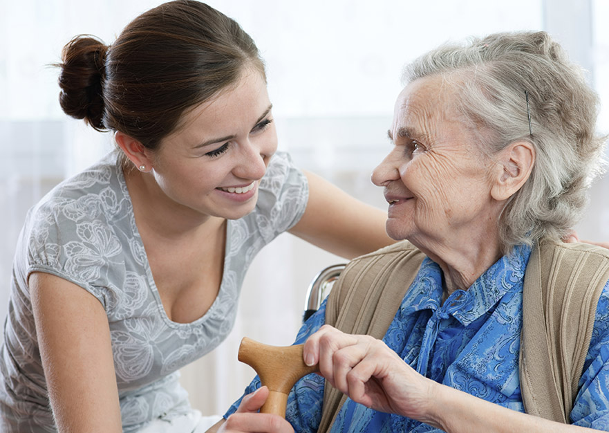 A young lady is smiling at the elderly woman she is assisting in a wheelchair. The older woman is also smiling back. 