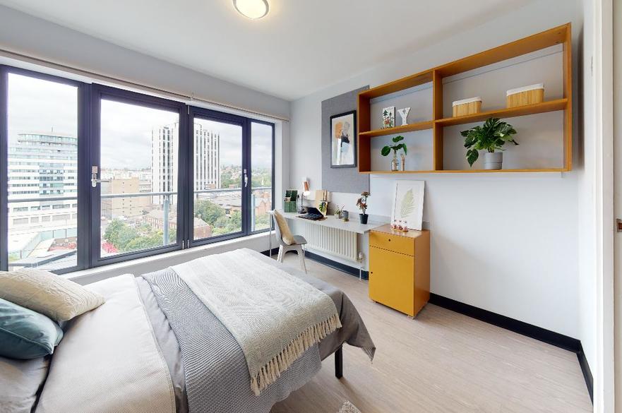 Room with white walls, a bed next to a window with a desk opposite and shelves on the wall above it. image