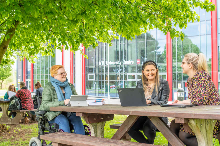 Three students working at a bench under a tree outside the Mansfield campus. One student is in a wheelchair. All are smiling in mid-conversation and using their laptops.