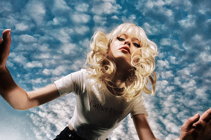 A young woman with blonde hair pictured from below against a blue sky.