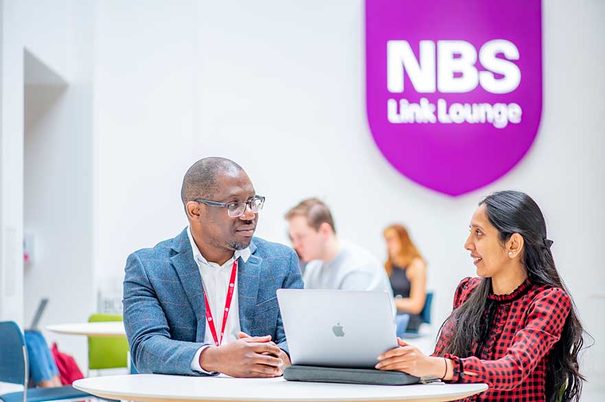 Male teacher and mature female student talking at a table with a purple Nottingham Business School sign in the background