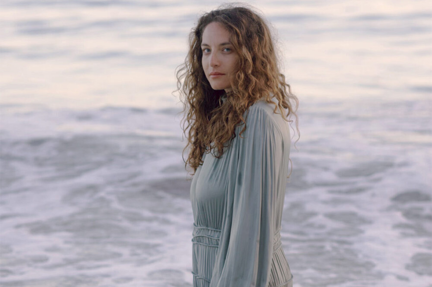 A woman with curly brown hair stood in the sea wearing a long grey dress.