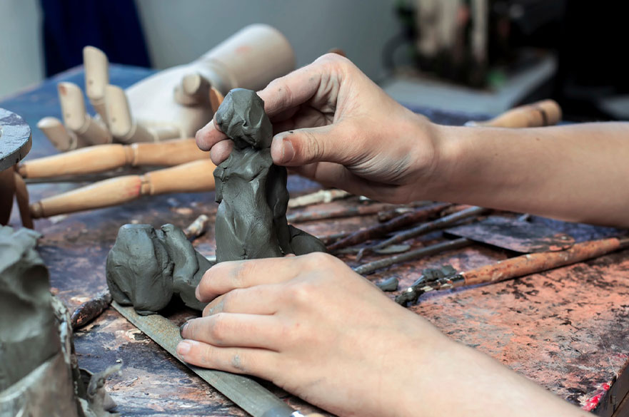 a pair of hands molding a figure out of clay on a messy workbench