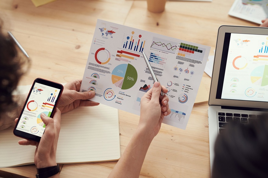 Person reviewing printed charts and graphs while viewing business analytics data on a smartphone and laptop.
