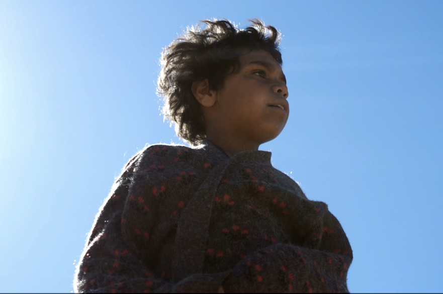 Still from the film In My Blood It Runs (2019). A young boy stands staring into the distance against a backdrop of clear blue sky.