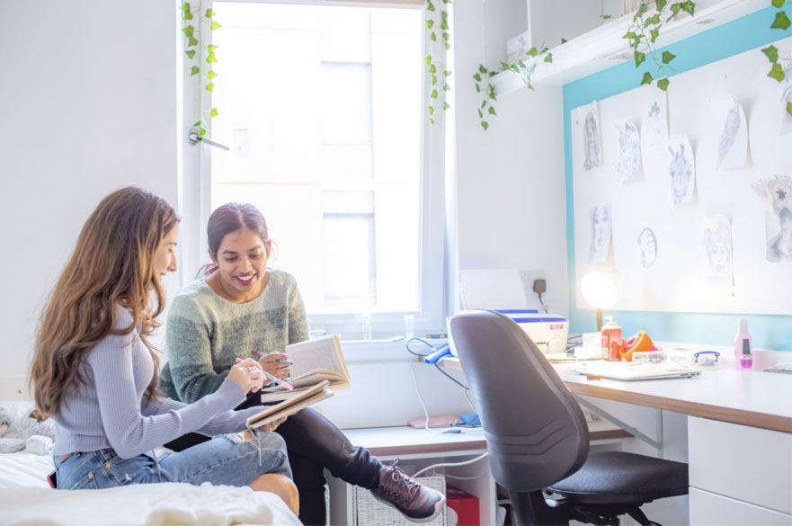 Two students sat on a bed in a brightly lit halls of residence.