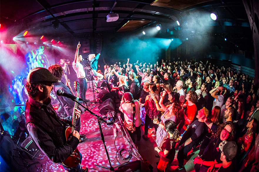 A band performing in front of a crowded audience taken from the side of the stage