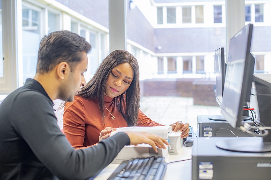 Two young people sat by computers looking through textbooks