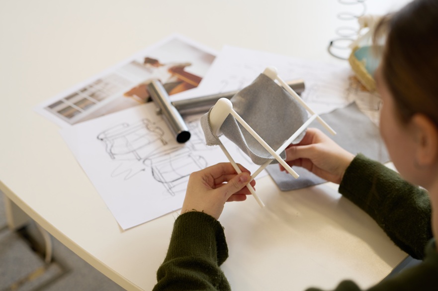 A student holding a scale model of her chair design with the design sketches on the table in front of her.