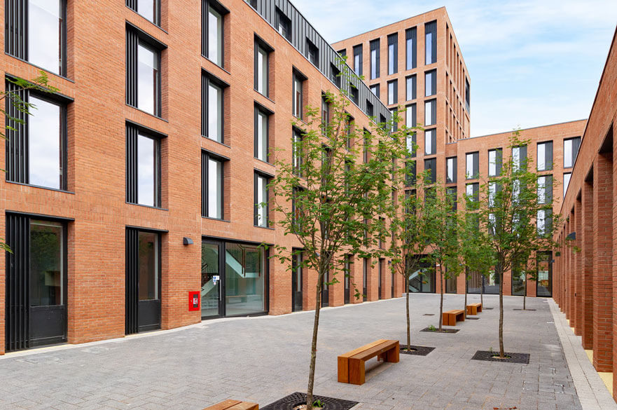 Exterior courtyard of a modern student accommodation building with brick fa&ccedil;ades, trees and seating areas.