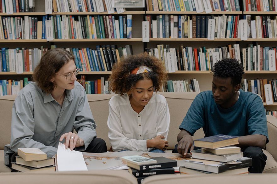 Three people sitting on a sofa reading books in front of a bookcase.