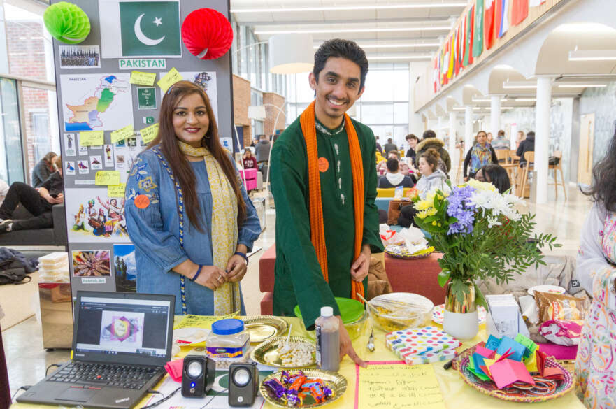 Two students stood at a stand displaying elements of Pakistani culture, with the flag of Pakistan behind them.