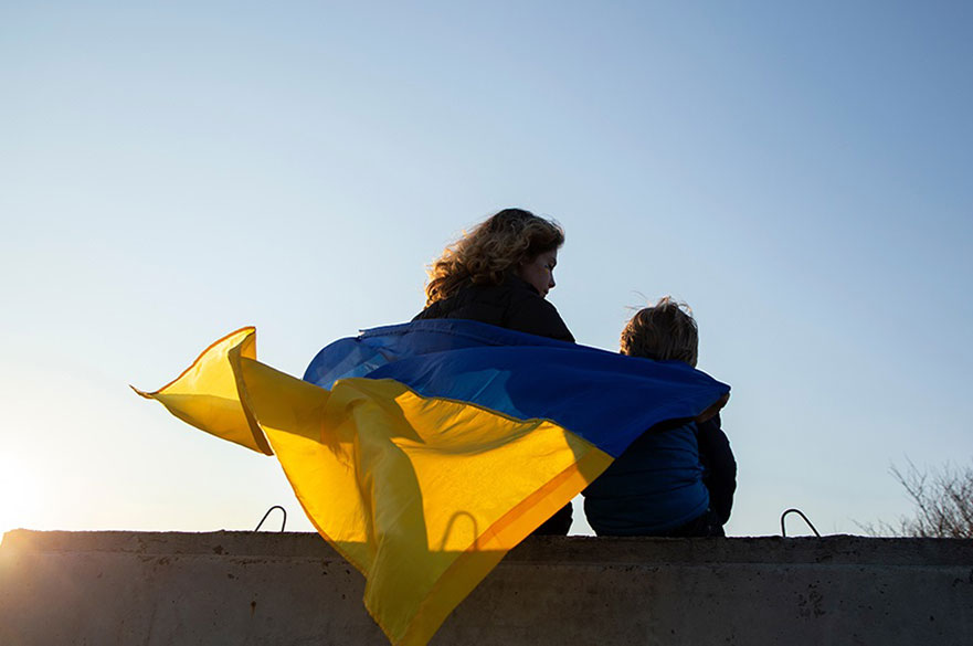 Little boy and a woman sit with their backs at sunset against the sky with a blue and yellow Ukrainian flag