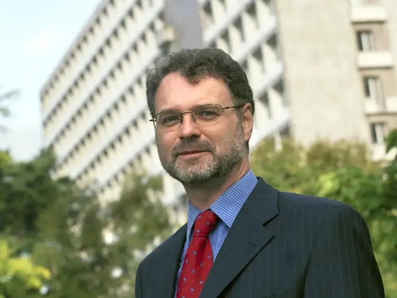 Person in a suit and red tie standing outdoors,  with a modern building and greenery in the background.