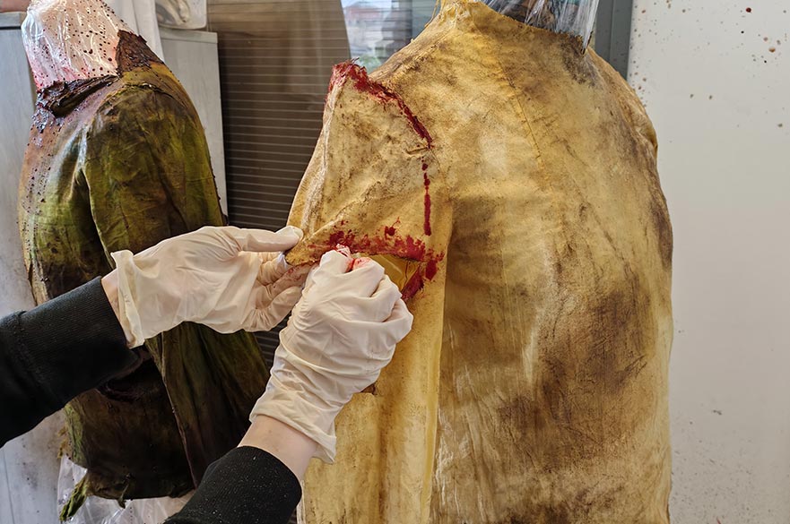 A student wearing protective gloves applies fake blood and dirt to the shoulder of a distressed costume jacket on a mannequin during a costume breakdown workshop. Another aged and weathered garment is visible in the background, showcasing techniques used in the Costume Making for Film and TV for 15–17 Year Olds short course at Nottingham Trent University.