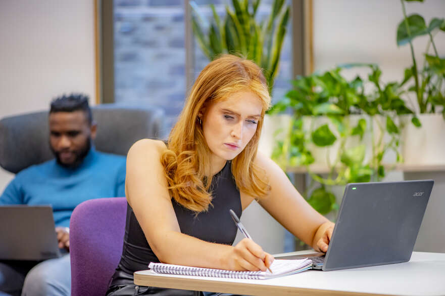 A student writing in a notepad and looking at a laptop