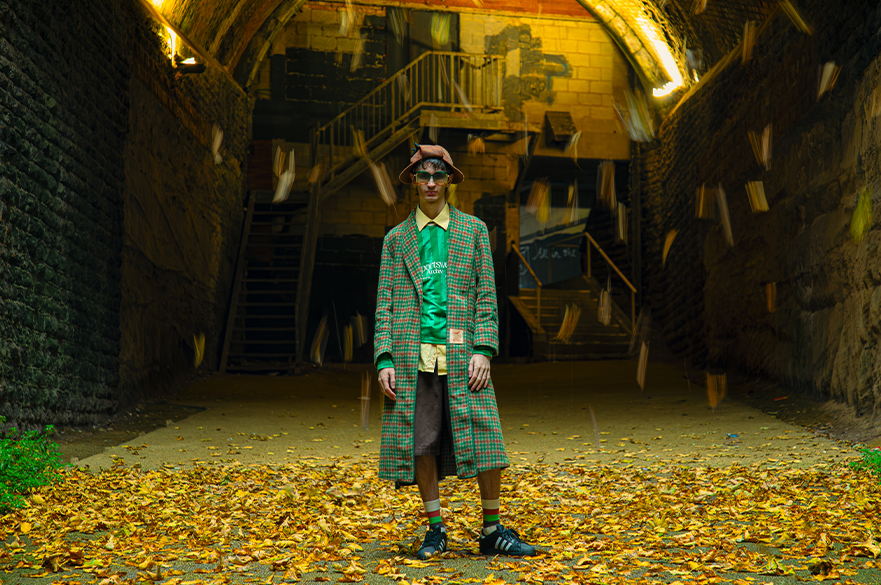 A model stands wearing a red hat, green shirt, shorts and green and red checked coat, with a backdrop of a derelict building under yellow lighting.
